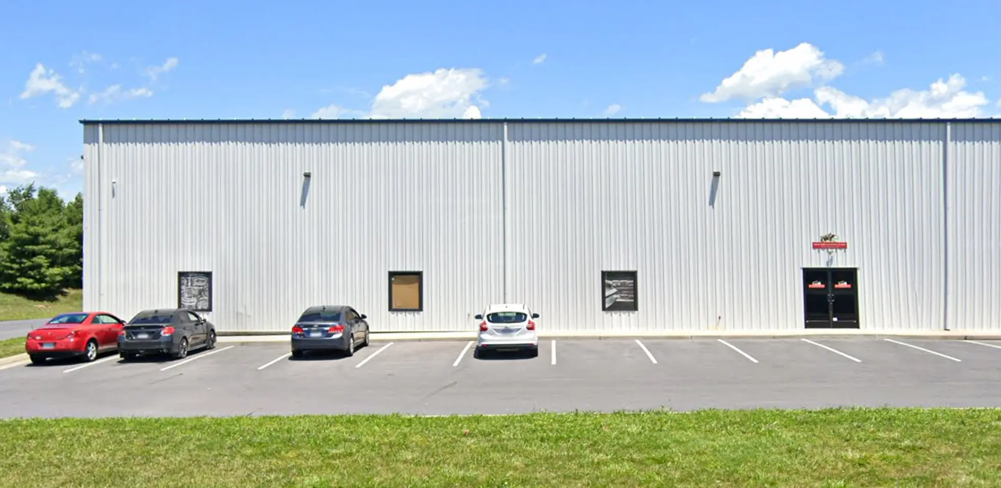 Industrial building with a metal facade, three parked cars in front, clear sky above, and grassy area in foreground. No recognizable landmarks present.