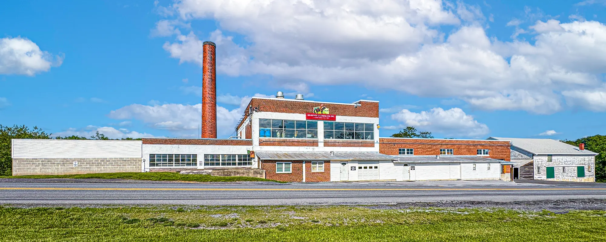 A large brick building with a smokestack, surrounded by grass, under a partly cloudy sky. A sign reads "The Hershey Company."