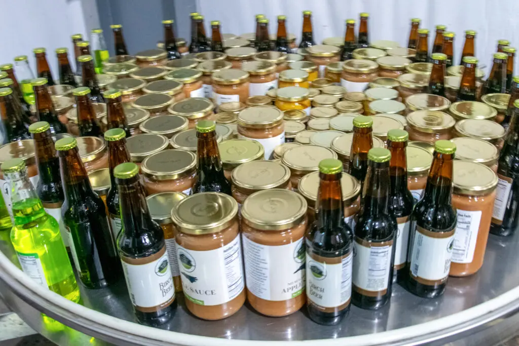Various jars and bottles are arranged on a round stainless steel table. Labels indicate sauces and beverages for sale or display.