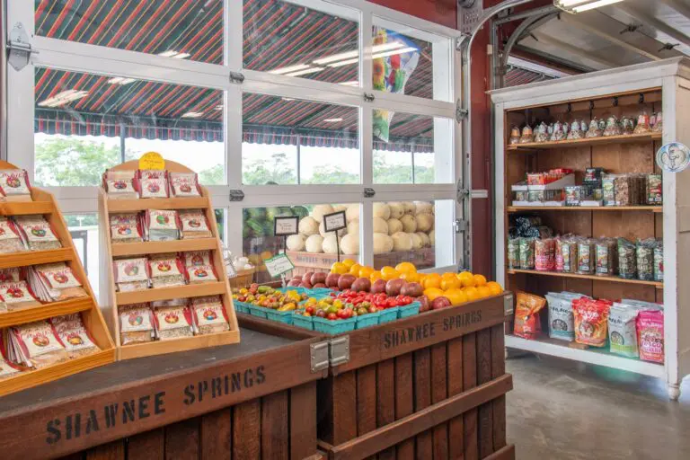 A quaint local market features fresh produce, packaged goods, and snacks. Wooden crates labeled "Shawnee Springs" decorate the colorful, inviting space.
