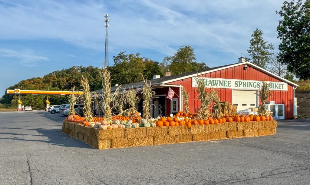 Red building labeled "SHAWNEE SPRINGS MARKET" surrounded by pumpkins and hay bales, located near a gas station and forested hill.