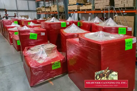 Stacks of red-wrapped pallets with "DO NOT STACK" signs are in a warehouse. A sign reads "Shawnee Canning Co. Inc." on the front pallet.