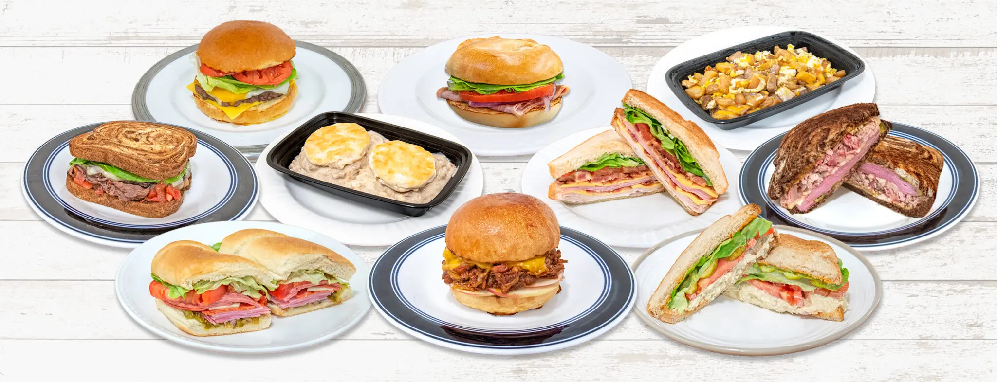 A variety of sandwiches and dishes are arranged on white plates, displayed on a light wooden surface for a food presentation.