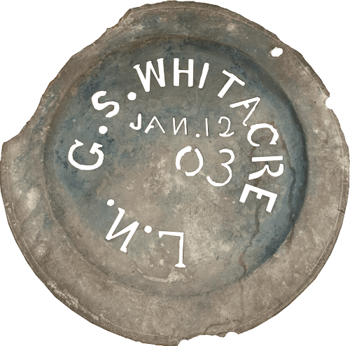 A round, weathered metal object features embossed text: "G.S. Whitacre, Jan. 12, 03," on its surface, hinting at historical significance.