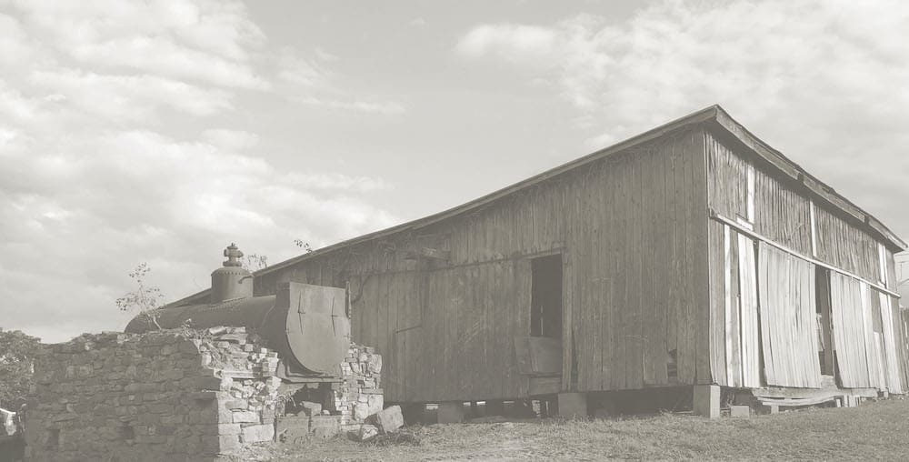 A vintage, weathered wooden barn stands beside a metal tank and stone remnants under a cloud-streaked sky, exuding rustic charm.