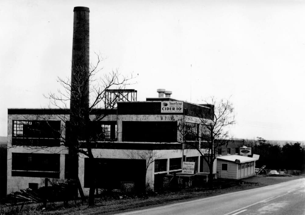 A vintage black-and-white photo captures a rustic cider company building by the roadside, with bare trees and a tall chimney.