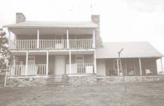 A two-story historic house with stone chimneys and a large porch, situated on a grassy lawn. No recognizable landmarks or persons visible.
