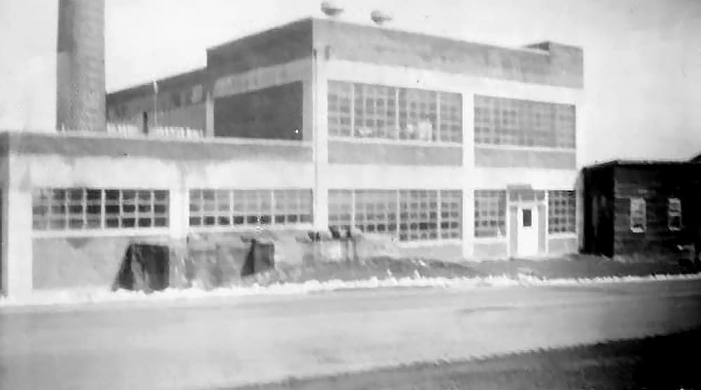 A monochrome image of a mid-20th-century industrial building with large windows, a smokestack, and adjacent small structure on a cleared street.