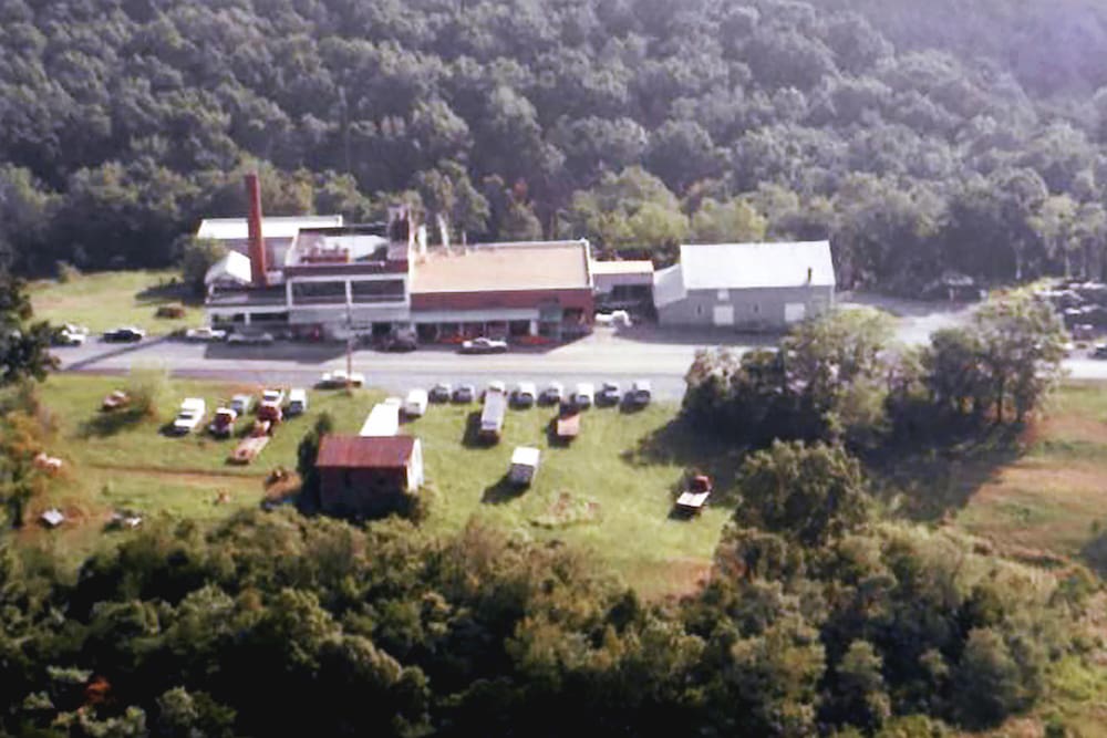 Aerial view of a rural factory surrounded by trees, with several parked vehicles and small buildings in a grassy area.