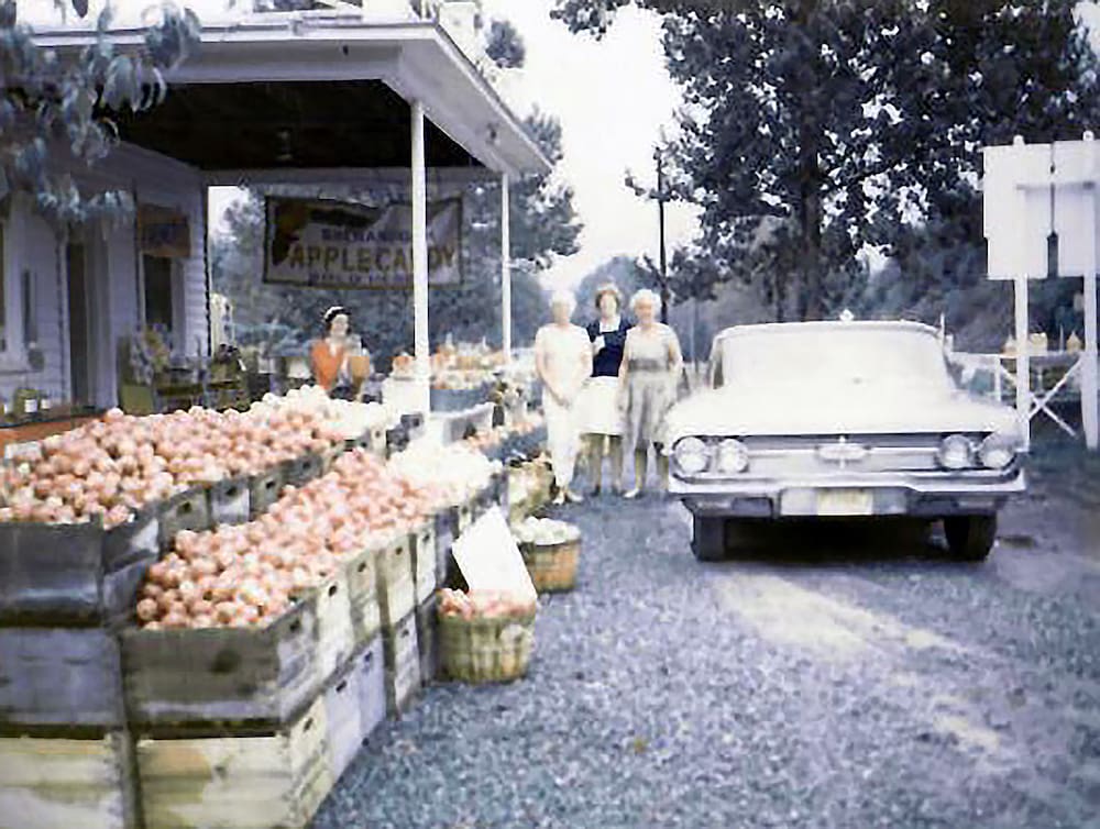 A roadside market with apples displayed in crates, a parked vintage car, and three people standing nearby in front of a white building.