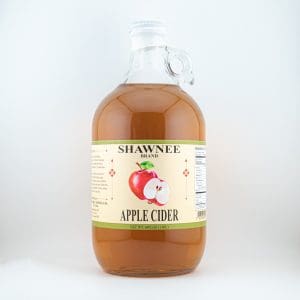 A clear jug filled with Shawnee brand apple cider, featuring a label with an apple graphic against a plain background.