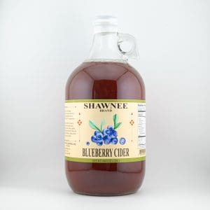 A glass jug with a handle contains Shawnee Brand Blueberry Cider, labeled with blueberry illustrations and nutritional information on a light yellow background.