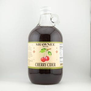 A clear glass jug of Shawnee Brand Cherry Cider, featuring a label with cherries and text, against a plain background.