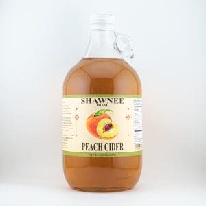 A clear jug of Shawnee Brand peach cider with a detailed label featuring an illustrated peach, positioned against a plain white background.