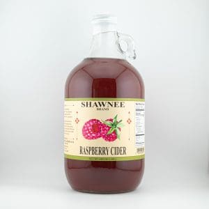 A clear glass jug of Shawnee Brand Raspberry Cider with a decorative label featuring raspberries, placed against a white background.