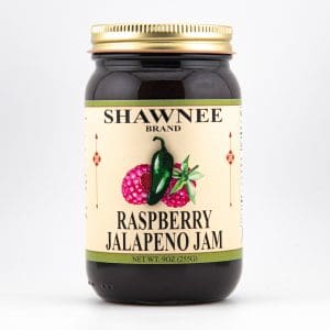 A jar labeled "Shawnee Brand Raspberry Jalapeno Jam," featuring illustrations of raspberries and a jalapeño, with a gold lid against a plain background.