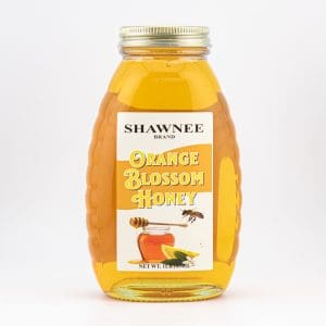 A jar of Shawnee Brand Orange Blossom Honey with a golden hue, decorative label, and metal lid is prominently displayed against a neutral background.