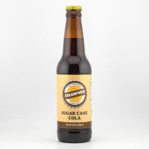 A glass bottle of Shawnee Sugar Cane Cola with a yellow label and brown cap, standing against a plain white background.