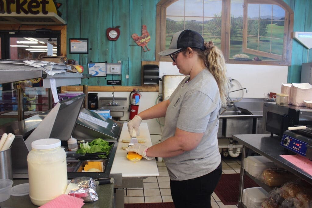 A person wearing a gray shirt and cap prepares sandwiches in a kitchen with teal walls, under a painting of a window view.