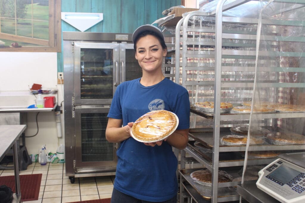 A person in a blue shirt and cap holds a pie in a bakery kitchen with racks of pastries and an industrial oven.