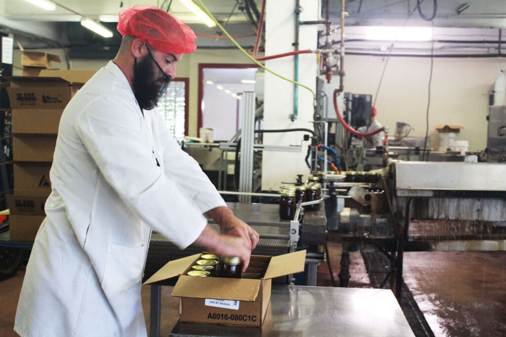 A person in a lab coat and hairnet packs jars in a busy industrial kitchen, with equipment and boxes around.