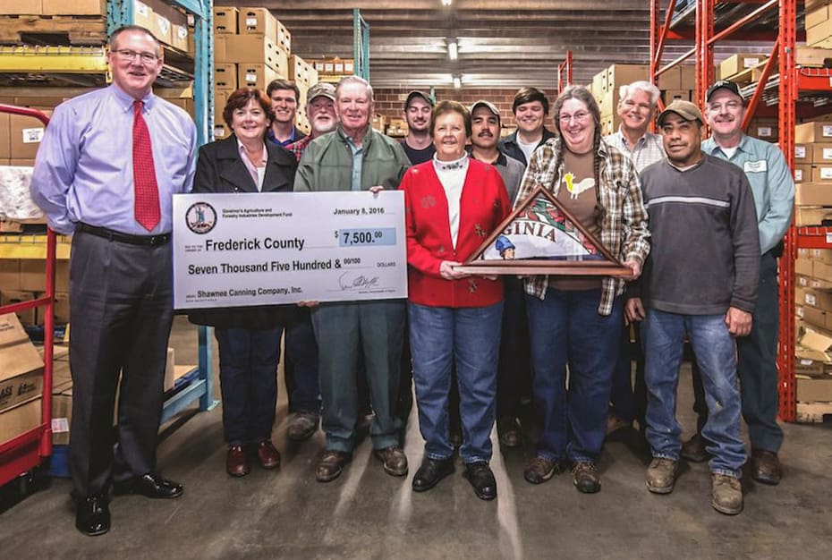 A group of people in a warehouse with a check and Virginia flag plaque, smiling for the camera. Shelves hold boxes in the background.